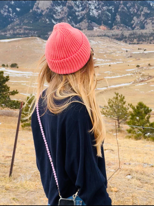 woman wearing a pink head and a crossbody pink beaded phone stripe looking at a field with trees and mountains in the back. 